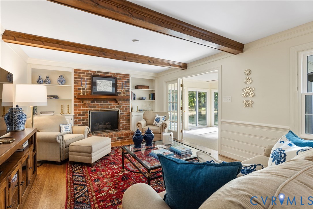 Exposed wood ceiling beams, brick fireplace with wood mantle, built-in shelving, wood-finish flooring, and French doors