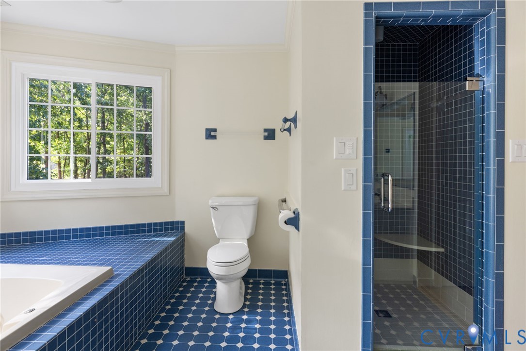 Bathroom with blue floor tiling, white window with grid panes, white toilet, tiled tub surround, and a glass-enclosed shower with blue tiling