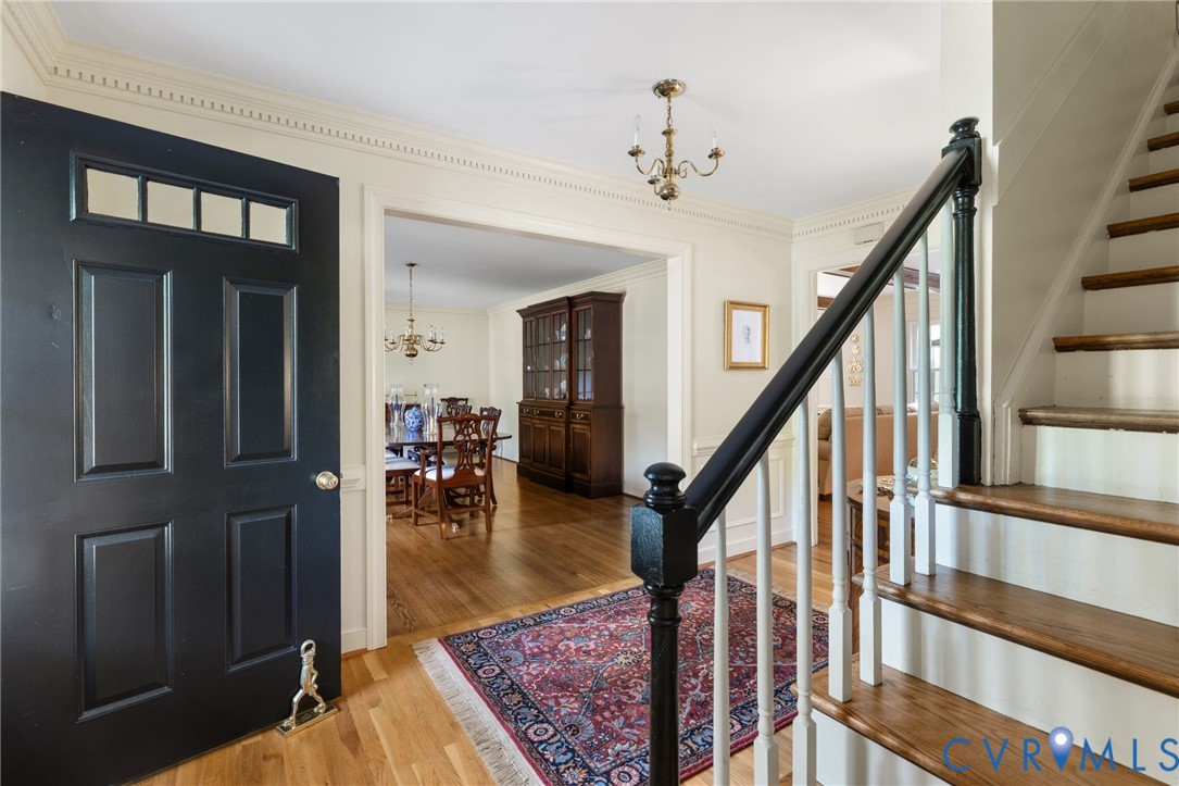 Black paneled entry door with transom windows, hardwood flooring, detailed crown molding, staircase with wood treads and balusters, and a brass chandelier