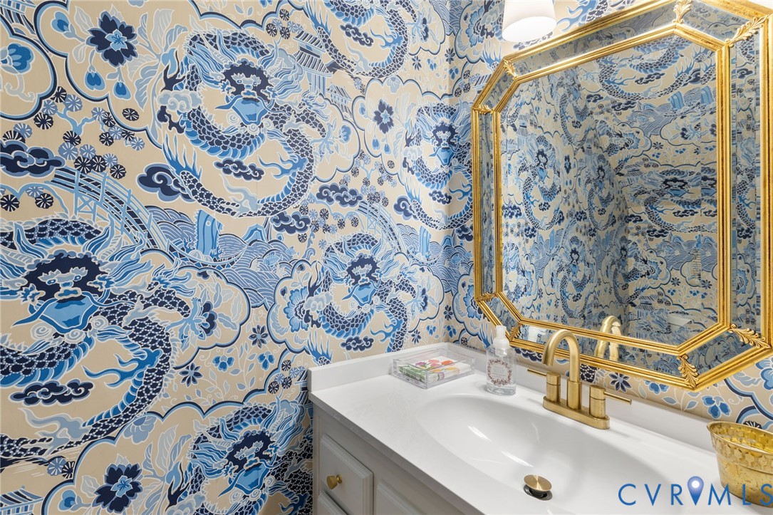 Powder room featuring decorative wallcovering, octagonal gold-framed mirror, white vanity with integrated sink, and brass-finish faucet