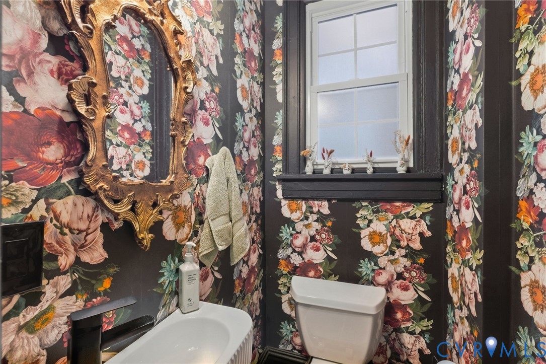 Decorative wallpaper, ornate gold-tone framed mirror, white single-hung window, dark window trim, and white toilet