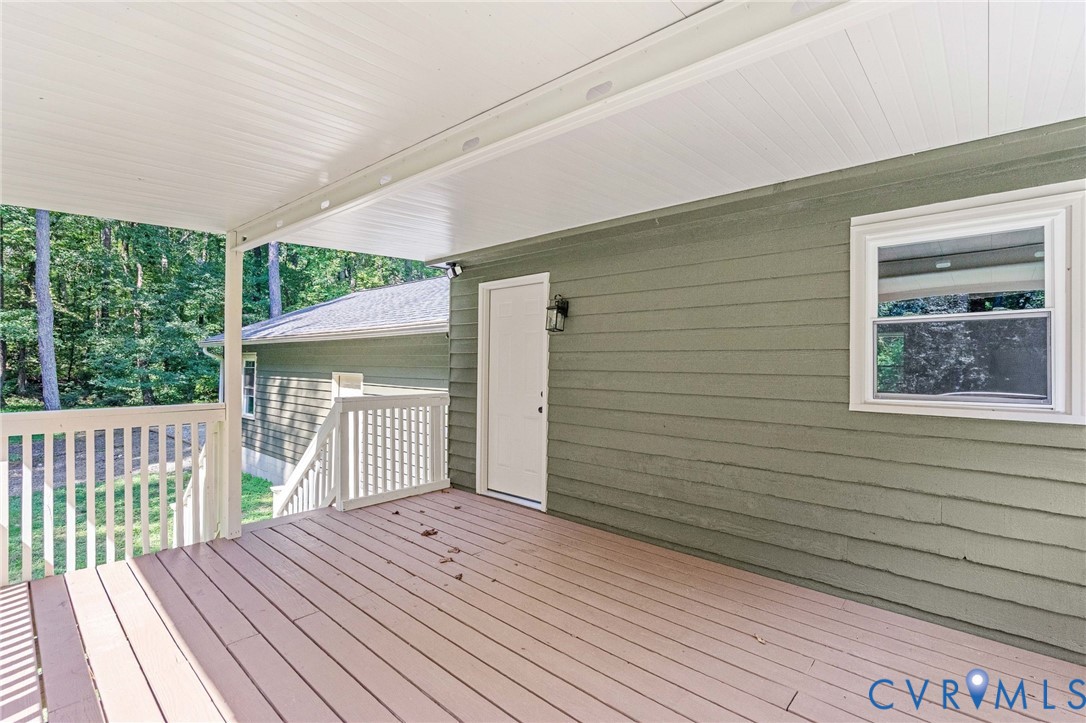 Covered outdoor deck with light wood-finish flooring, white railing, and a door with an exterior light fixture