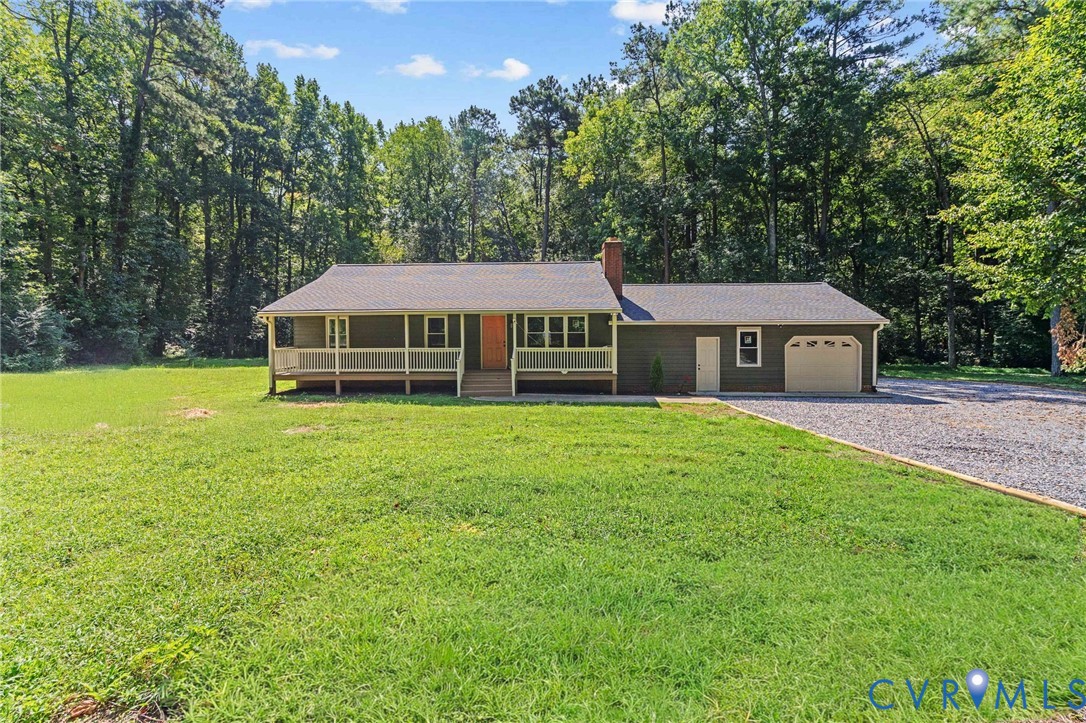Ranch-style exterior with a covered front porch, attached garage, and gravel driveway, set against a wooded backdrop