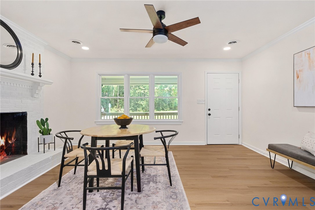 Hardwood-style flooring throughout, white painted brick fireplace with mantel, crown molding, three-panel window, and ceiling fan with integrated lighting