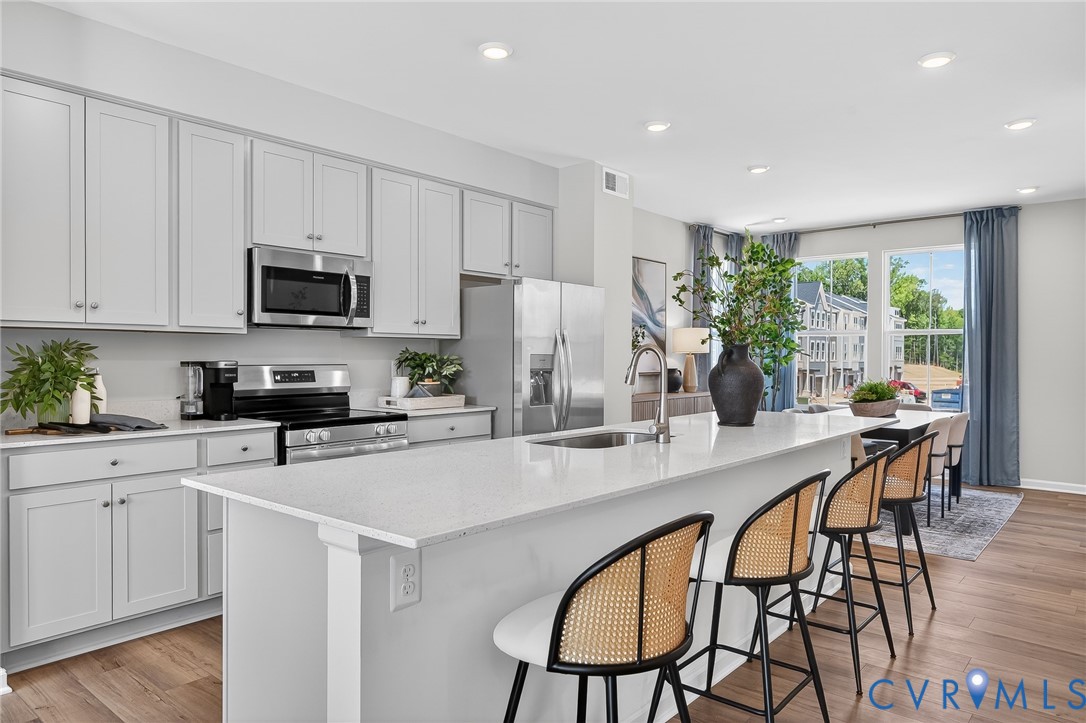Kitchen featuring light-toned cabinetry, stainless steel appliances, a center island with sink, quartz countertops, and wood-finish flooring