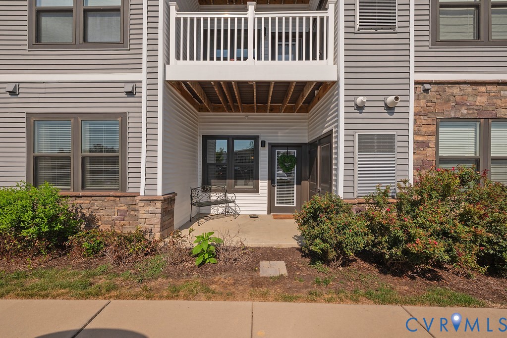 Ground-level entry with a covered patio, framed by stone veneer accents and a white railing balcony above Ground-level entry with a covered patio, framed by stone veneer accents and a white railing balcony above