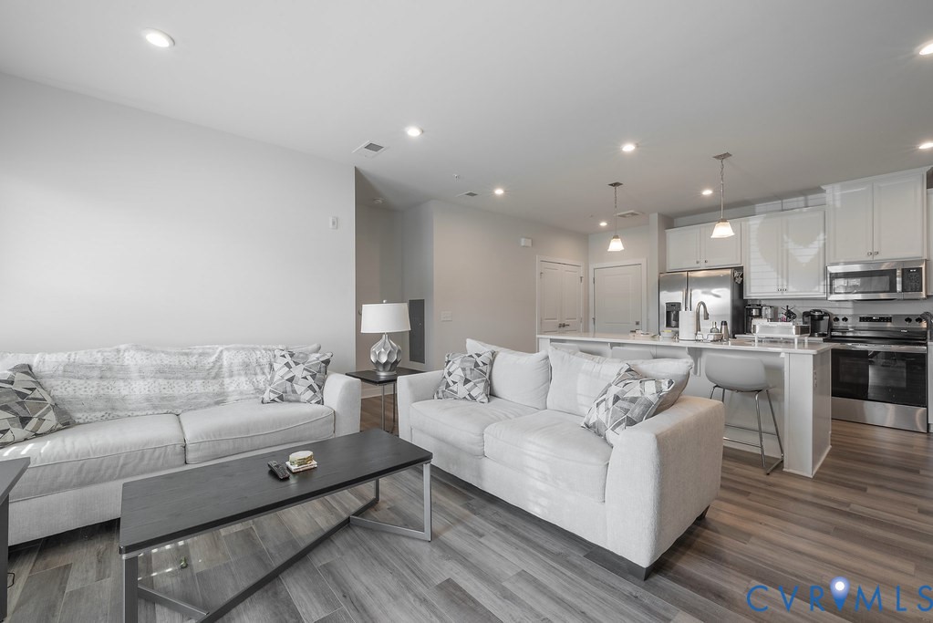 Open-concept living space featuring wood-finish flooring, recessed lighting, and a kitchen with white cabinetry, stainless steel appliances, and a breakfast bar Open-concept living space featuring wood-finish flooring, recessed lighting, and a kitchen with white cabinetry, stainless steel appliances, and a breakfast bar