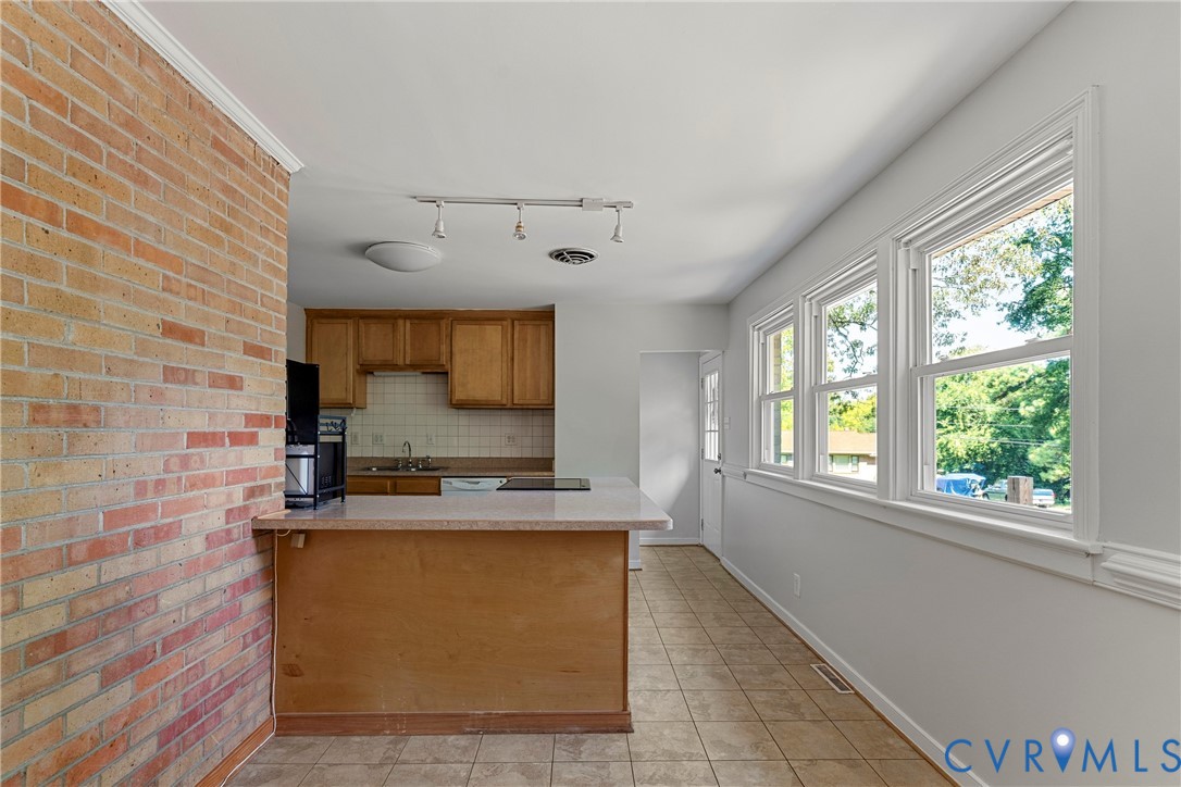 Brick accent wall, L-shaped kitchen counter, light wood-finish cabinetry, tile flooring, and a wall of windows