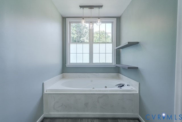 Soaking tub with a white surround and integrated faucet, under a window with privacy film, illuminated by a multi-pendant light fixture, complemented by two floating shelves