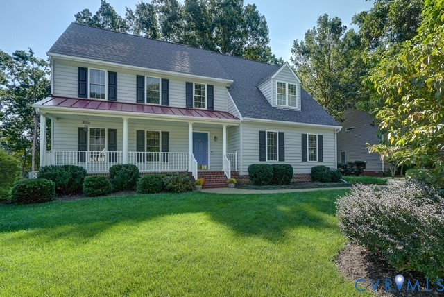 Traditional two-story residence featuring light-colored siding, dark shutters, a covered front porch with white railings, a prominent dormer window, and mature landscaping