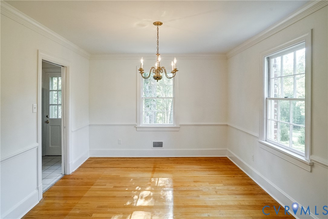 Sunny dining room with hardwood flooring, two large windows, brass chandelier, crown molding, and wainscoting