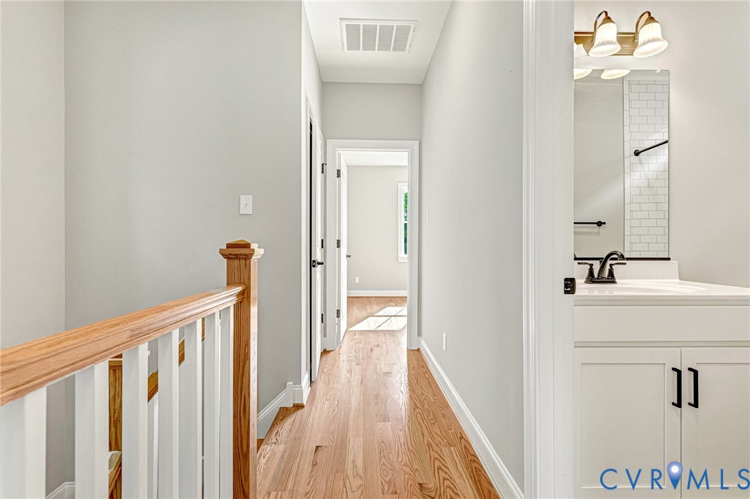 Hardwood flooring in the hallway, white painted walls, wood railing with white spindles, white vanity with a dark faucet, subway tile backsplash in the bathroom