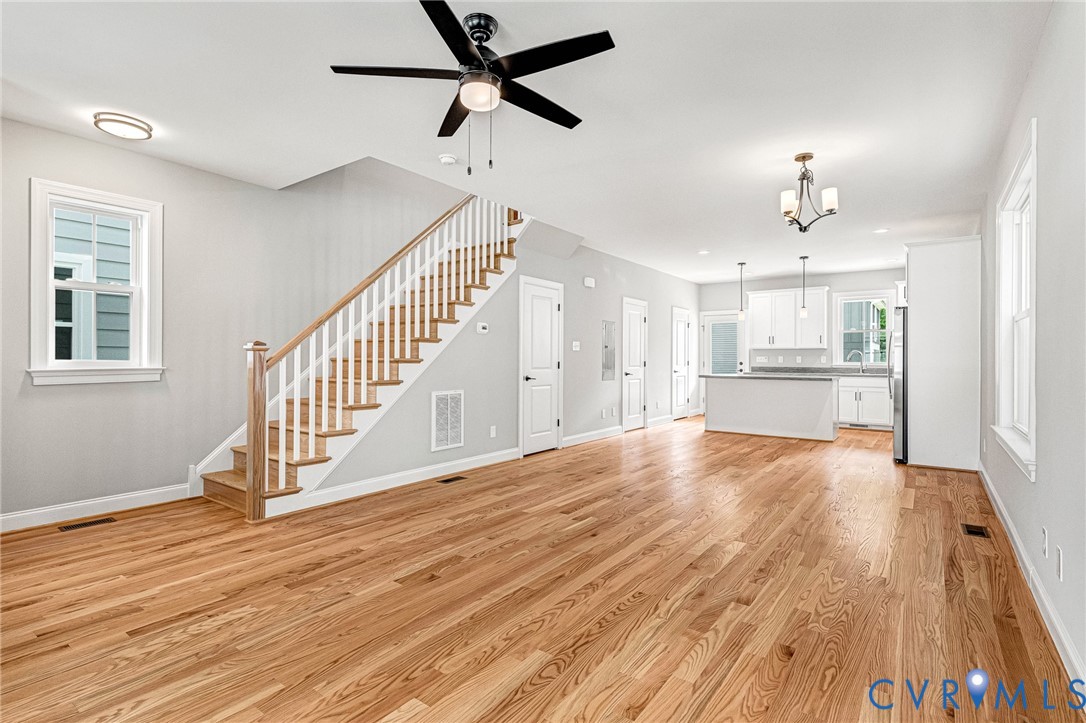 Open-concept interior featuring wood-finish flooring, a white-spindled staircase with wood handrails, and a contemporary kitchen with white cabinetry and an island Open-concept interior featuring wood-finish flooring, a white-spindled staircase with wood handrails, and a contemporary kitchen with white cabinetry and an island