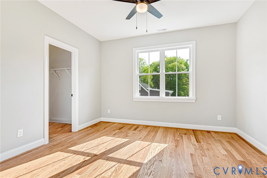 Bright, sunlit bedroom featuring hardwood flooring, large double-hung windows with natural light, ceiling fan with integrated lighting, and a walk-in closet with shelving and hanging rod