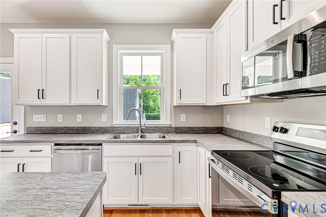 White shaker-style cabinetry with matte black hardware, stainless steel appliances, a double basin sink, light-toned countertop, and wood-finish flooring