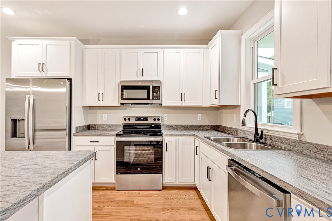 Modern kitchen featuring white shaker cabinetry, stainless steel appliances, grey stone-patterned countertops, wood-finish flooring, and a double basin sink with a matte black faucet