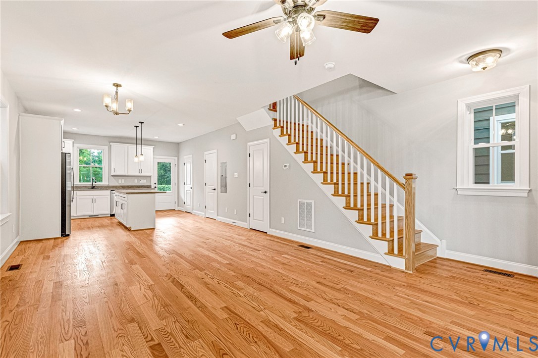 Hardwood flooring throughout, a staircase with wooden treads and white risers, a kitchen island with pendant lighting, white kitchen cabinetry, and an attached ceiling fan with lighting