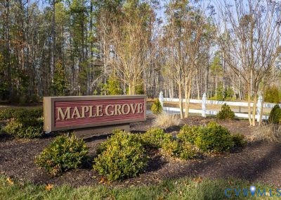 Maple Grove community signage featuring a red and tan sign, surrounded by landscaped shrubbery and trees, with a white fence in the background