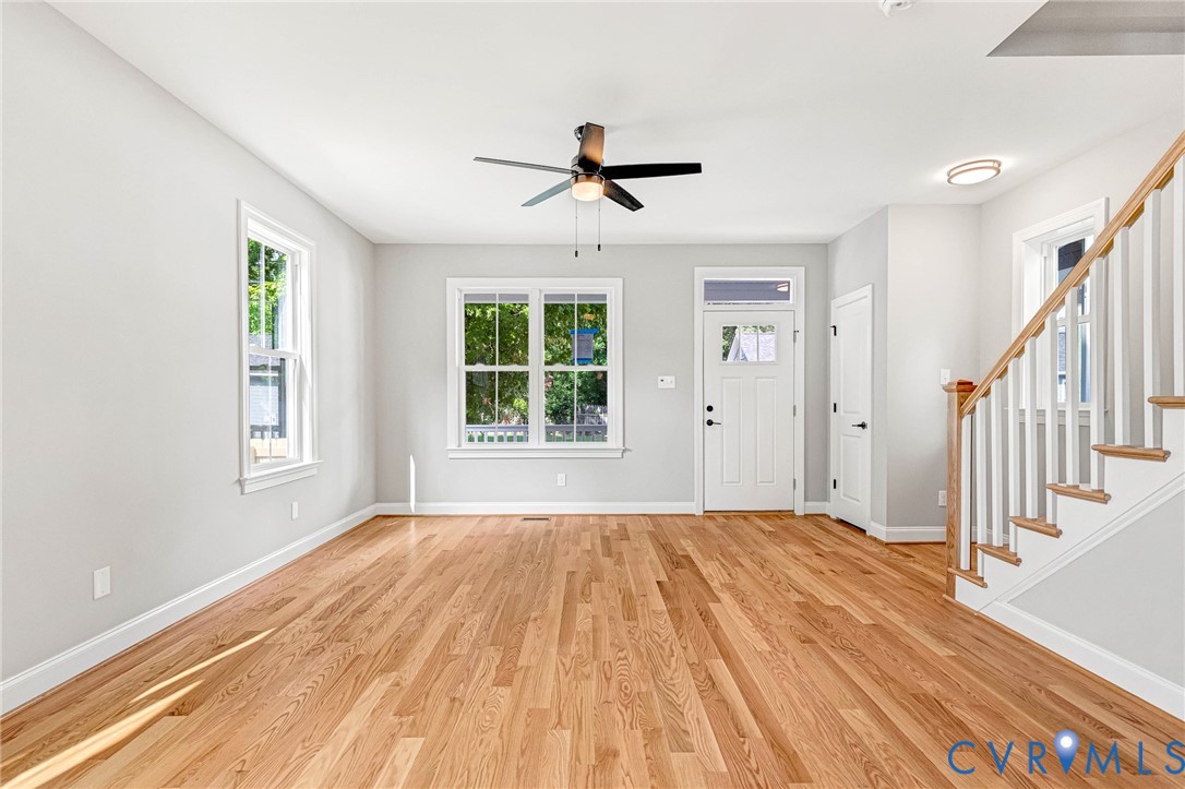 Hardwood flooring, multiple windows, a ceiling fan, a front entry door with a transom, and a staircase with white balusters Hardwood flooring, multiple windows, a ceiling fan, a front entry door with a transom, and a staircase with white balusters