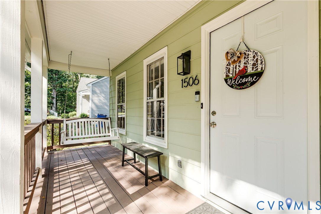 Covered front porch featuring wood-finish flooring, white porch swing, white entry door, and exterior wall lantern
