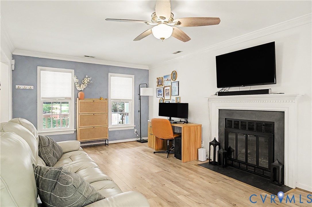 Wood-finish flooring, ceiling fan with integrated lighting, fireplace with a black insert and white mantel, and multiple windows