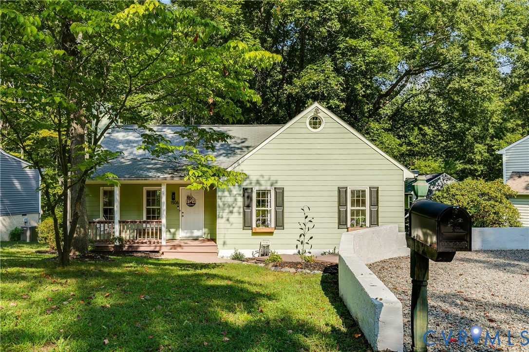 Light green siding with dark shutters, front porch with wood railings, a white front door, and a gravel driveway