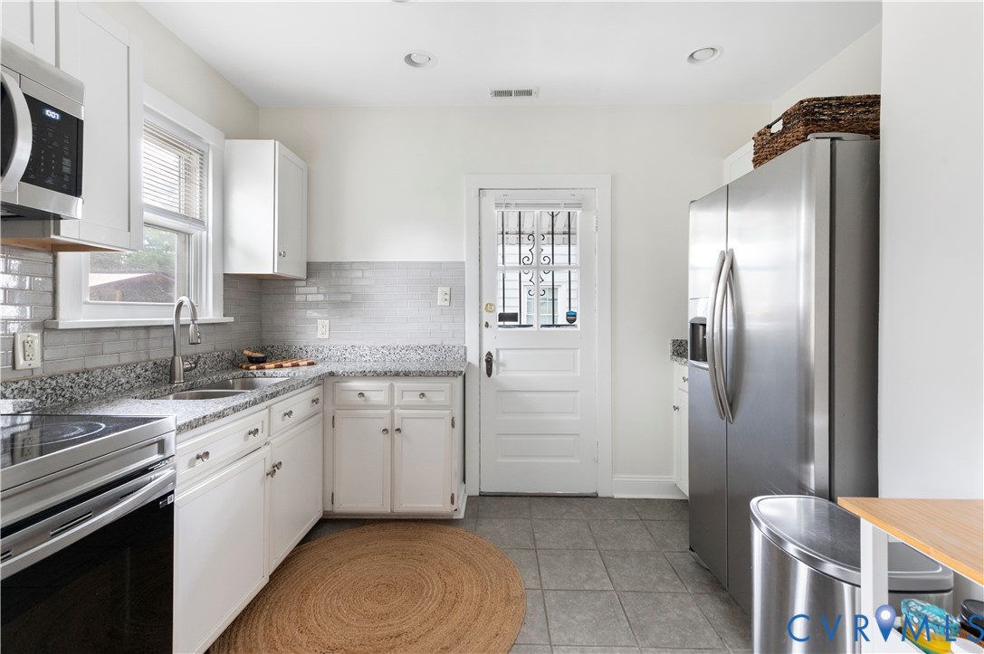 White cabinetry with dark hardware, granite countertops, tile backsplash, built-in stainless steel microwave, and stainless steel refrigerator White cabinetry with dark hardware, granite countertops, tile backsplash, built-in stainless steel microwave, and stainless steel refrigerator