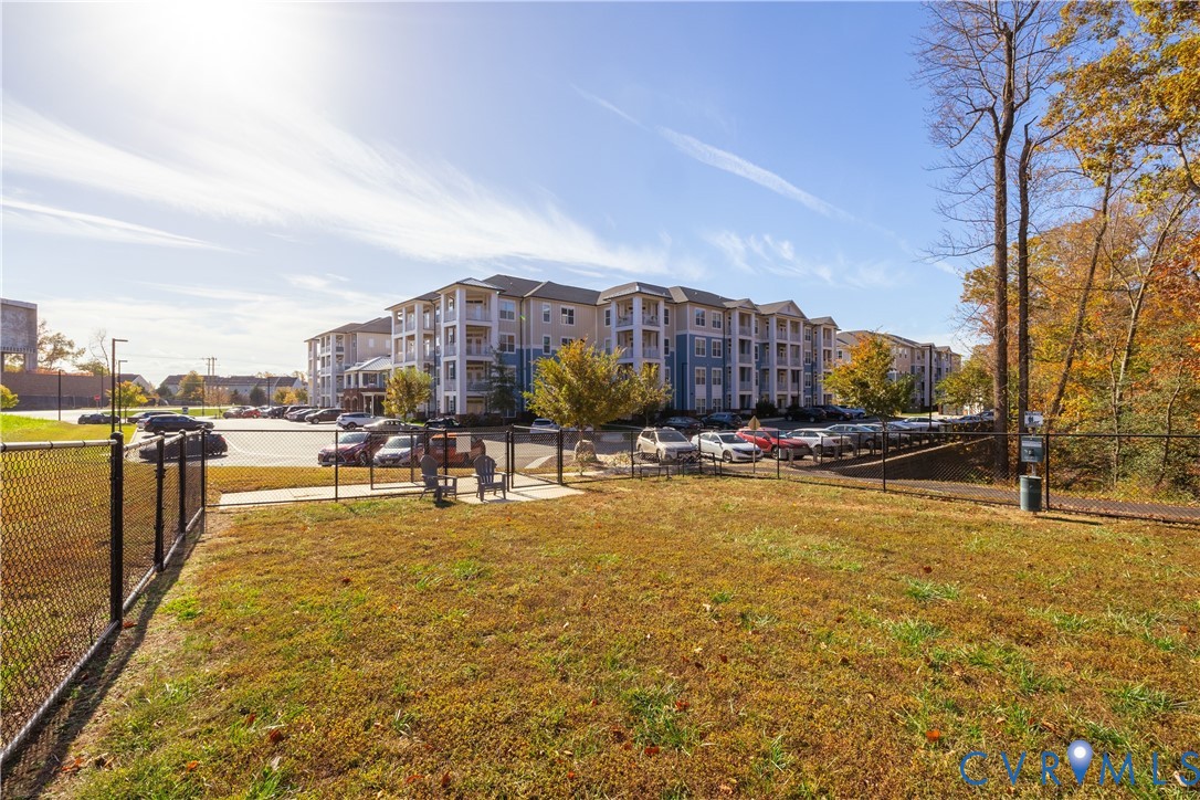 Fenced green space with a paved walking path, adjacent to a parking area and multi-story residential buildings