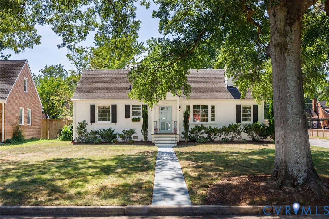 White facade with dark shutters, light blue front door, front yard with mature trees, and a paved walkway