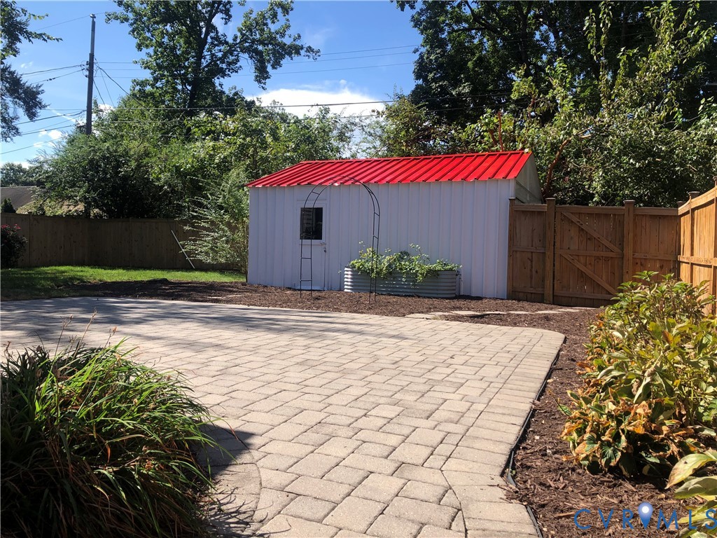 Paver patio with a decorative border, a white storage shed with a red metal roof, garden beds, and wood privacy fencing.