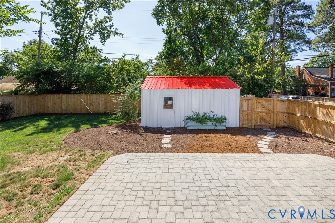 Paver patio with a stone pathway, wood privacy fencing, a storage shed with a red metal roof, and a raised garden bed