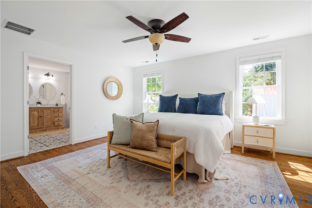 Hardwood flooring throughout, ceiling fan with integrated lighting, multiple windows, and a bathroom entry featuring a double vanity with round mirrors and wall-mounted lighting