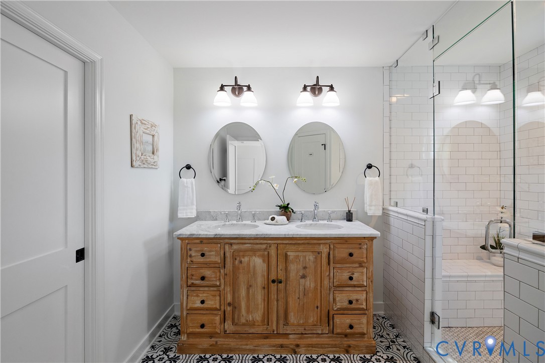 Dual vanity with a stone countertop, individual oval mirrors, and a glass-enclosed shower featuring subway tile