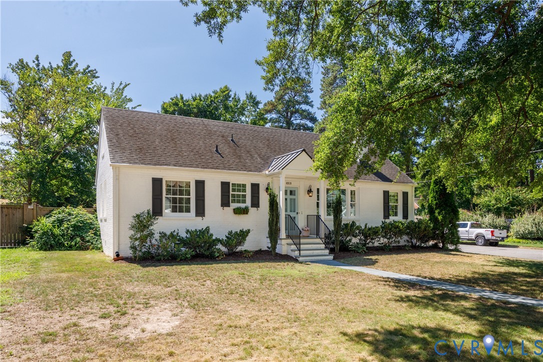 White brick exterior with dark shutters, gabled roofline, front porch with black railings, and a paved driveway