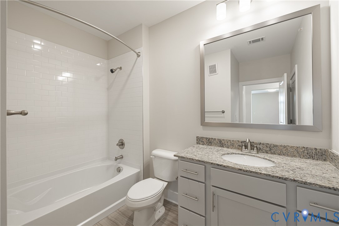 Bathroom featuring a white subway tile shower surround, a bathtub with brushed nickel fixtures, a single vanity with a stone-finish countertop, and wood-finish flooring