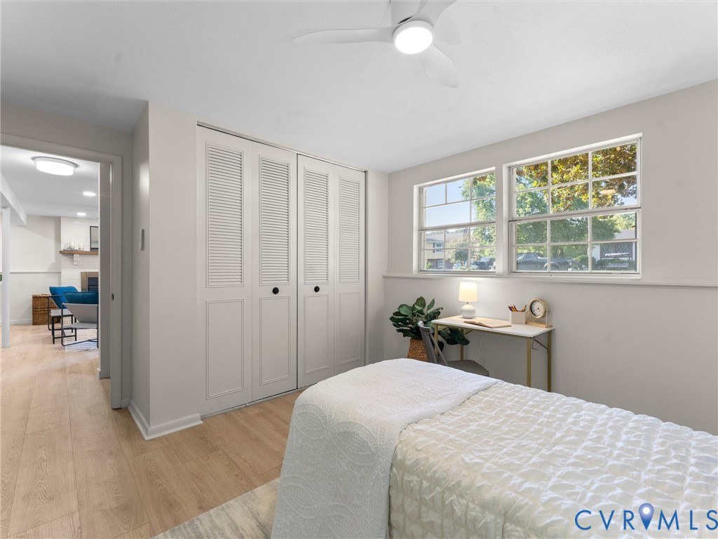 Light wood-finish flooring, two large windows, white louvered bifold closet doors, and an overhead ceiling fan with integrated lighting