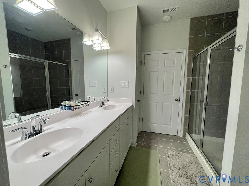 Dual vanity with undermount sinks and chrome fixtures, expansive wall mirror, tile flooring, glass-enclosed shower with tile surround, and overhead lighting