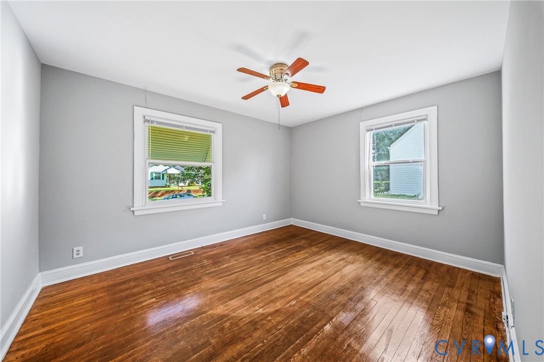 Wood flooring, light gray wall paint, two windows with white trim, and a ceiling fan with wood-tone blades