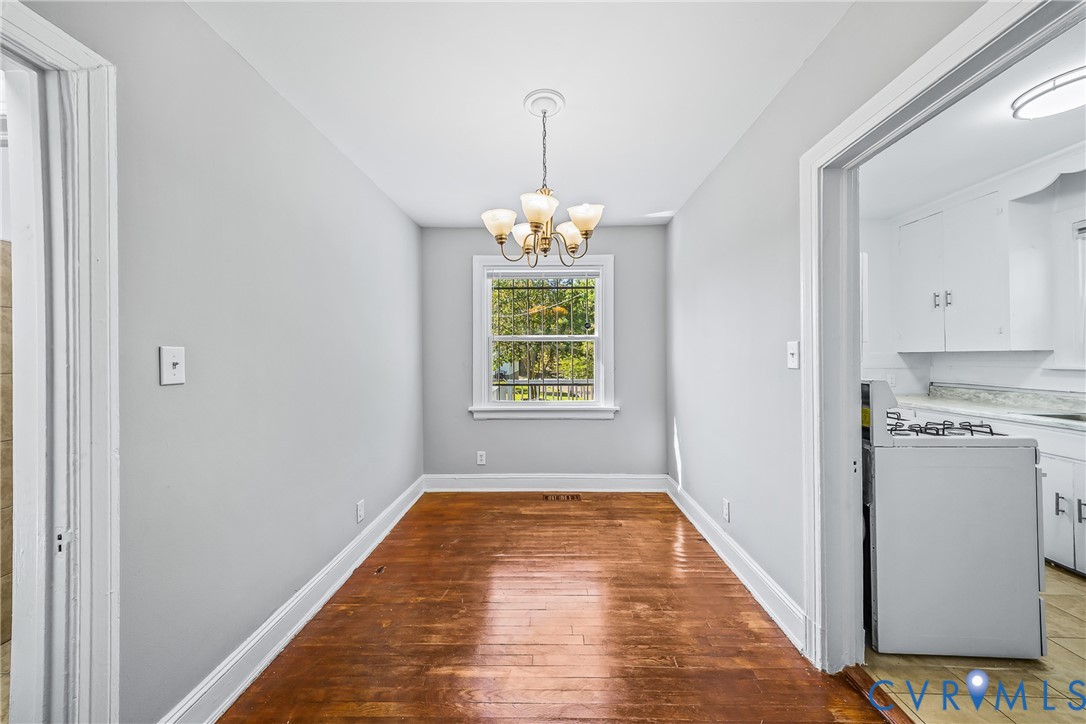 Hardwood flooring, central window, ceiling-mounted chandelier, white trim, and a doorway to a kitchen area