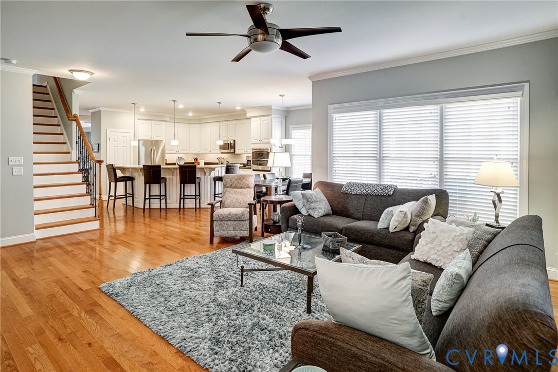 Hardwood flooring throughout, a staircase with wood treads and a wrought iron baluster, a kitchen with white cabinetry and an integrated island, and a ceiling fan with dark wood-finish blades