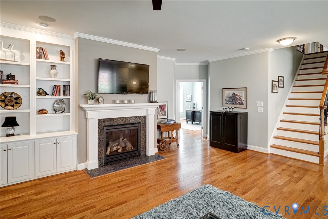 Hardwood flooring throughout, built-in cabinetry with shelving, fireplace with a stone surround and hearth, and a staircase with wood treads and a wood railing
