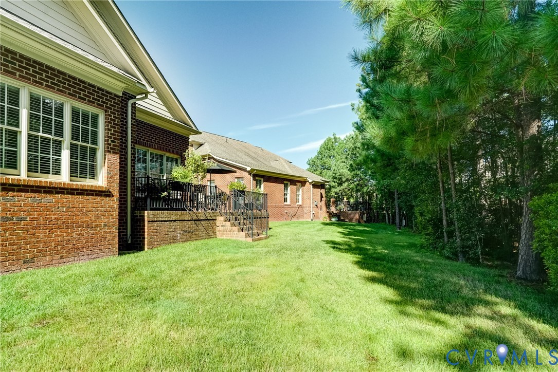 Expansive lawn with mature trees and a brick exterior, featuring multiple windows with louvered shutters and a raised patio with metal railings