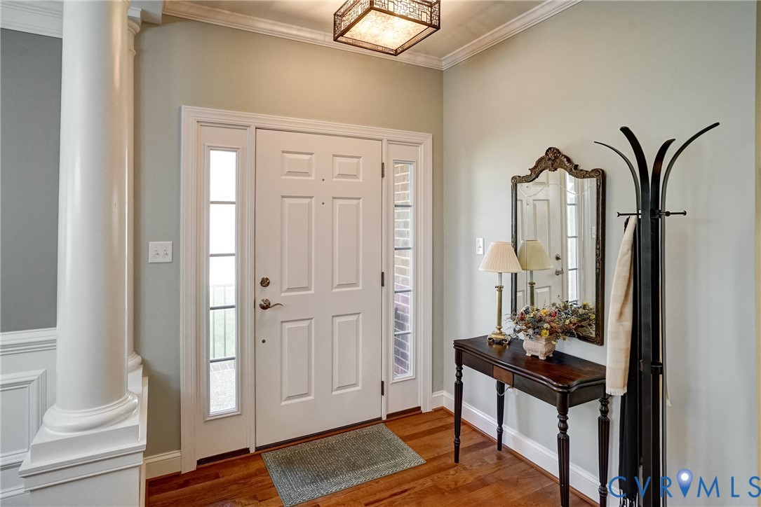 Solid wood flooring, paneled front door with sidelights, decorative ceiling light fixture, white crown molding, and wainscoting