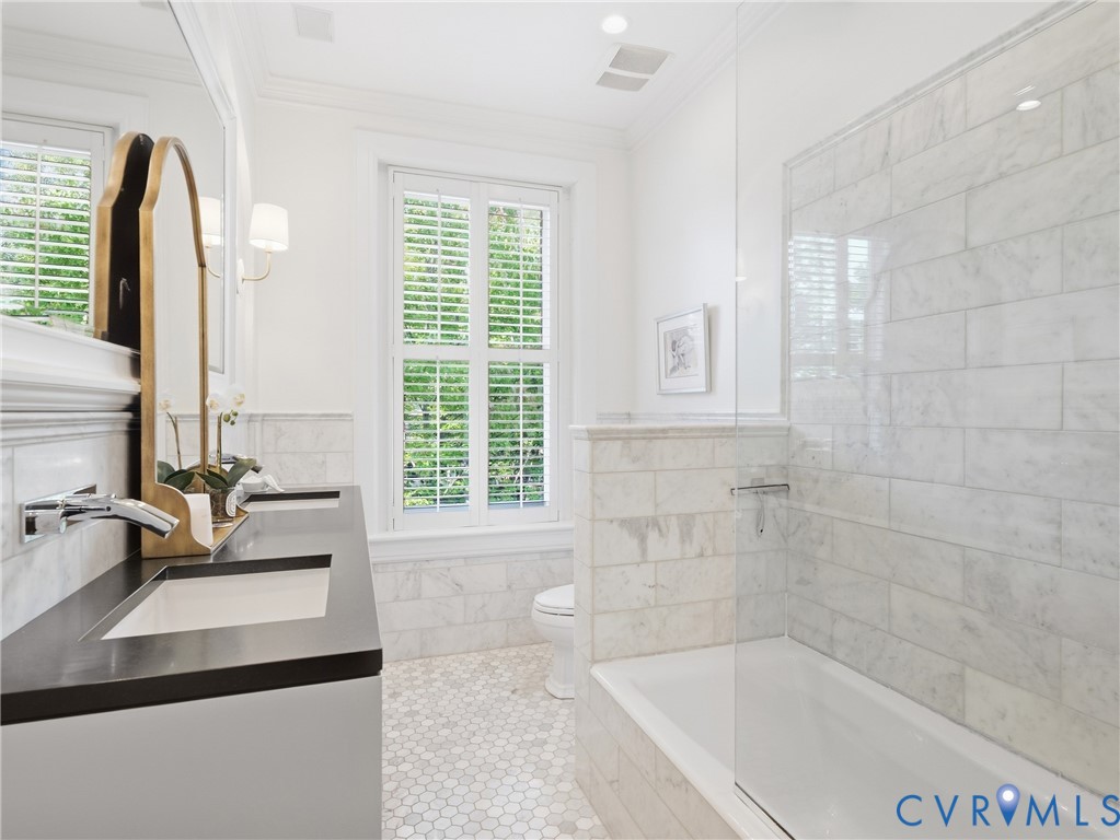 Double vanity with integrated sinks and dark countertops, white hexagon marble flooring, white subway tile bath surround, built-in bathtub, and plantation shutters