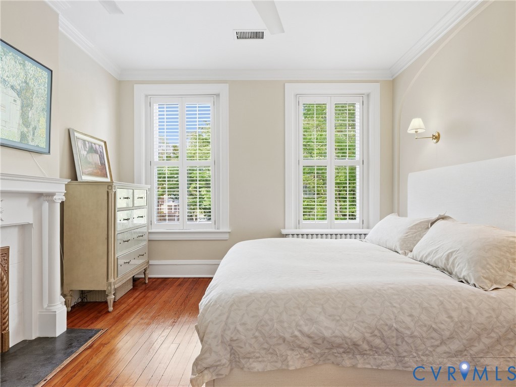 Hardwood flooring, crown molding, double windows with plantation shutters, wall-mounted sconce, and fireplace with mantel