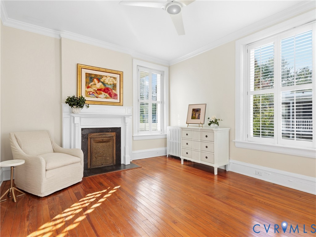 Hardwood flooring with a decorative fireplace and white mantel, crown molding, and multiple windows with plantation shutters