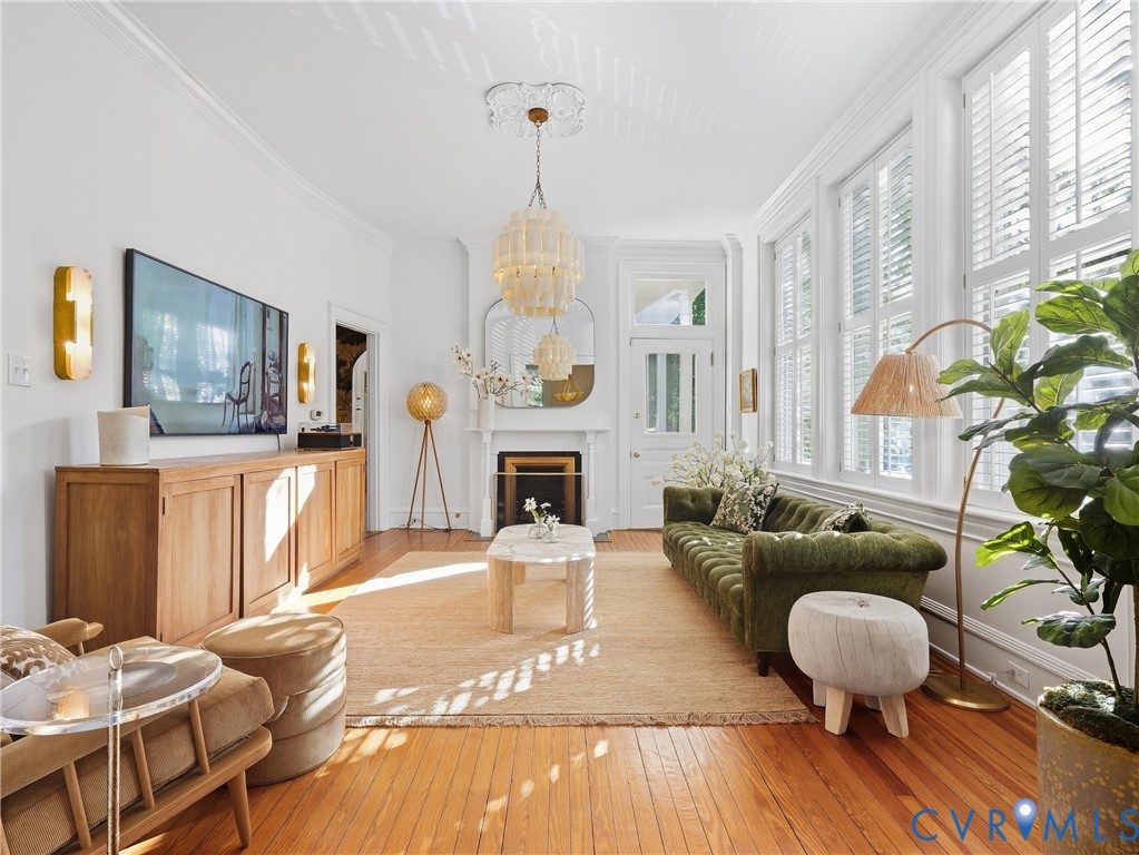Warm-toned wood flooring, multiple tall windows with plantation shutters, crown molding, a decorative fireplace, and a light-toned chandelier