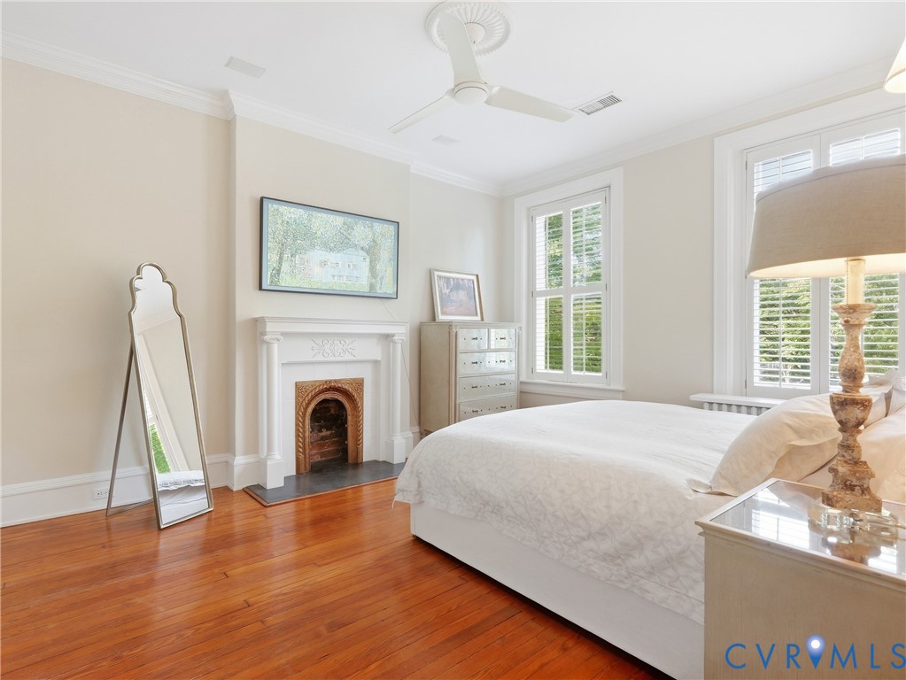 Hardwood flooring, fireplace with decorative mantel, crown molding, ceiling fan, and multiple windows with plantation shutters