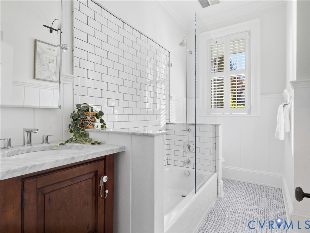 White subway tile shower surround, glass shower enclosure, wood-finish vanity with countertop, plantation shutters, and patterned floor tiling