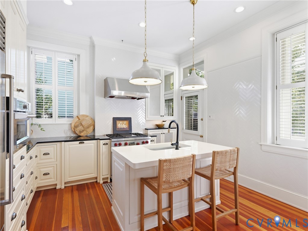 Hardwood flooring, central island with an integrated sink, built-in stainless steel appliances, white cabinetry, and a herringbone tile backsplash